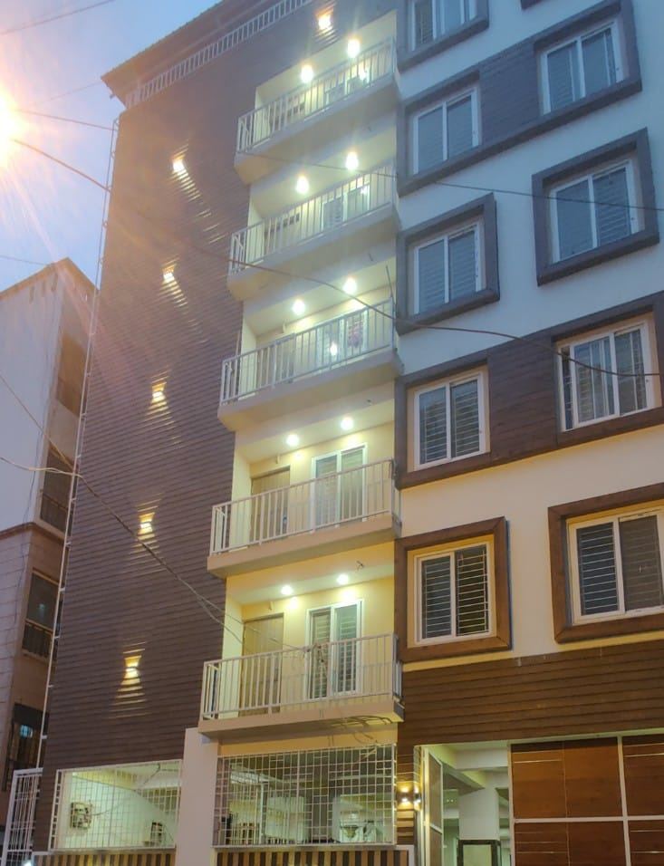 Nova Regency lit at dusk from the road — six floors of balconies, most of them lit, above the timber-clad stair core