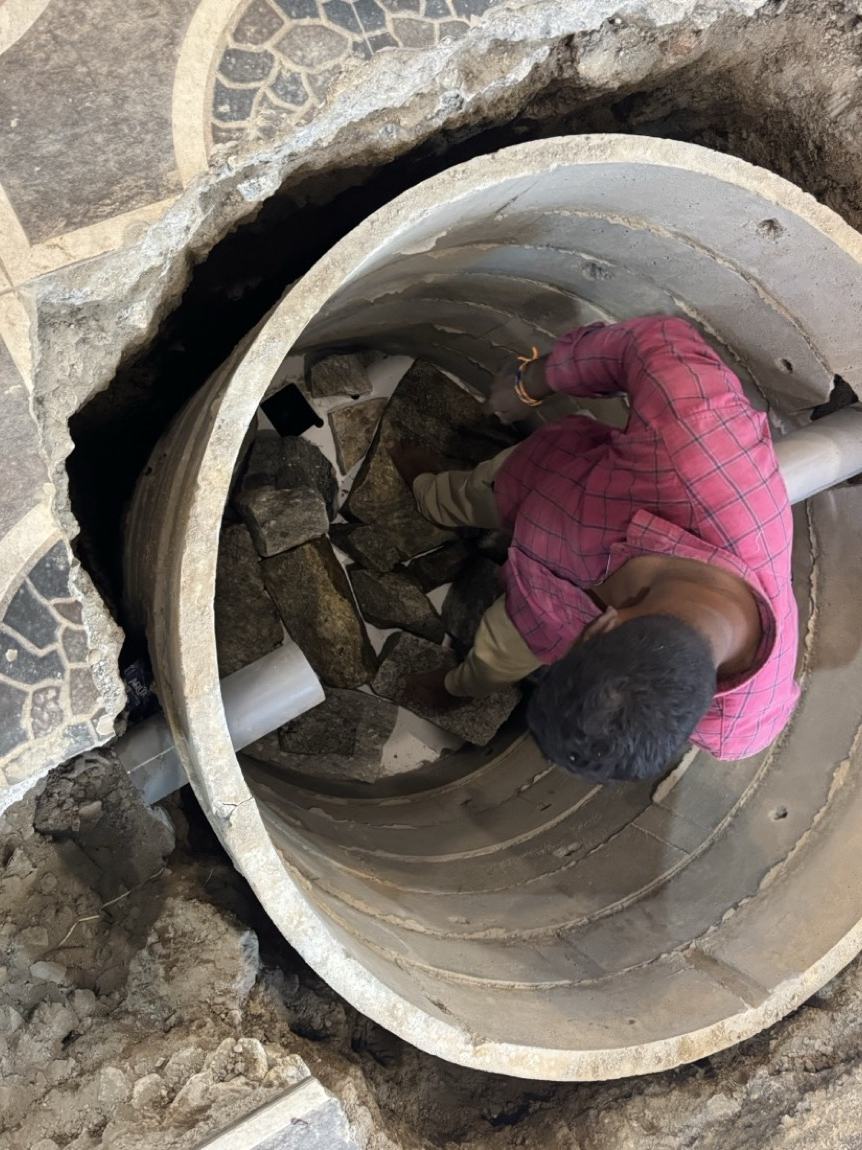 A worker inside the concrete chamber hand-packing broken stone around the inlet pipe