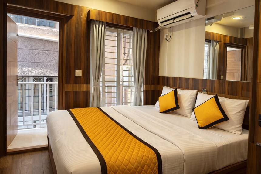A Deluxe 1BHK — the bed beside a full-height balcony window, daylight across the room